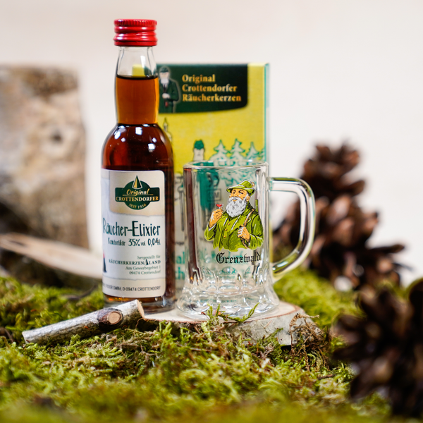 Das Geschenkset Grenzwald von Original Crottendorfer mit einer Kräuter-Räucher-Elixier-Flasche, einem bärtigen Glaskrug und Erzgebirgischer Weihnachtsduft-Räucherkerzen ruht auf grünem Moos inmitten von Tannenzapfen und Holzstäbchen.