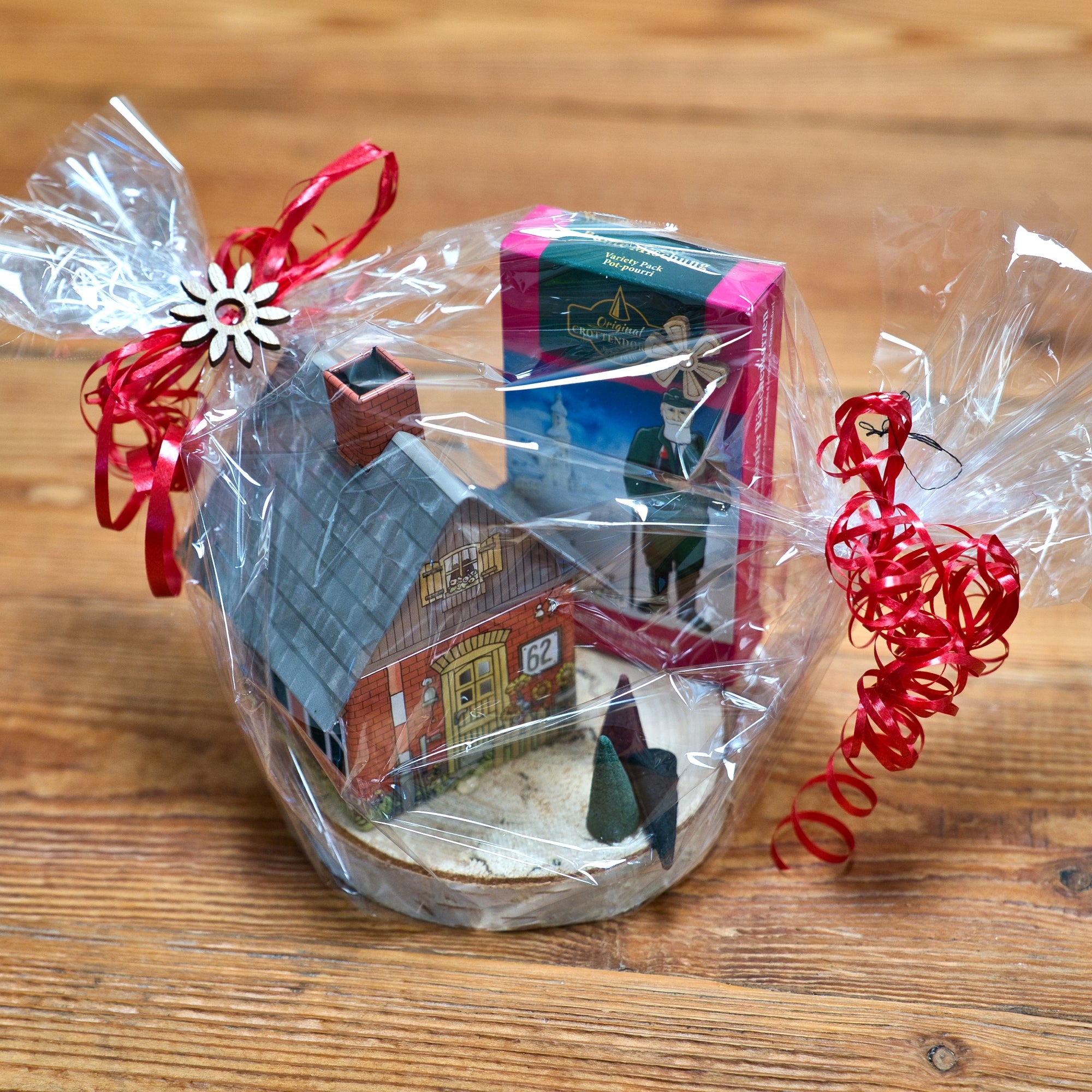 Das Geschenkset Räucherhäuschen + Räucherkerzen von Original Crottendorfer Räucherkerzen, bestehend aus einem charmanten Hausornament und einer Schachtel mit herrlichen Räucherkerzen, liegt auf einer Holzoberfläche. Es ist in transparente Folie mit einer roten Schleife eingewickelt und mit einer kleinen blumenförmigen Dekoration geschmückt.
