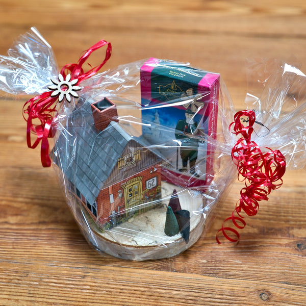 Das Geschenkset Räucherhäuschen + Räucherkerzen von Original Crottendorfer Räucherkerzen, bestehend aus einem charmanten Hausornament und einer Schachtel mit herrlichen Räucherkerzen, liegt auf einer Holzoberfläche. Es ist in transparente Folie mit einer roten Schleife eingewickelt und mit einer kleinen blumenförmigen Dekoration geschmückt.