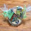 Ein in transparentes Zellophan gewickelter Geschenkkorb mit grünen Bändern enthält das Original Crottendorfer Räucherkerzen-Geschenkset mit Miniatur-Schachteln in Hausform, die an charmante Räucherhäuschen erinnern, einer gelben Schachtel mit Sonnenblumendekor aus Holz und kleinen kegelförmigen Räucherkerzen auf einer Holzoberfläche.