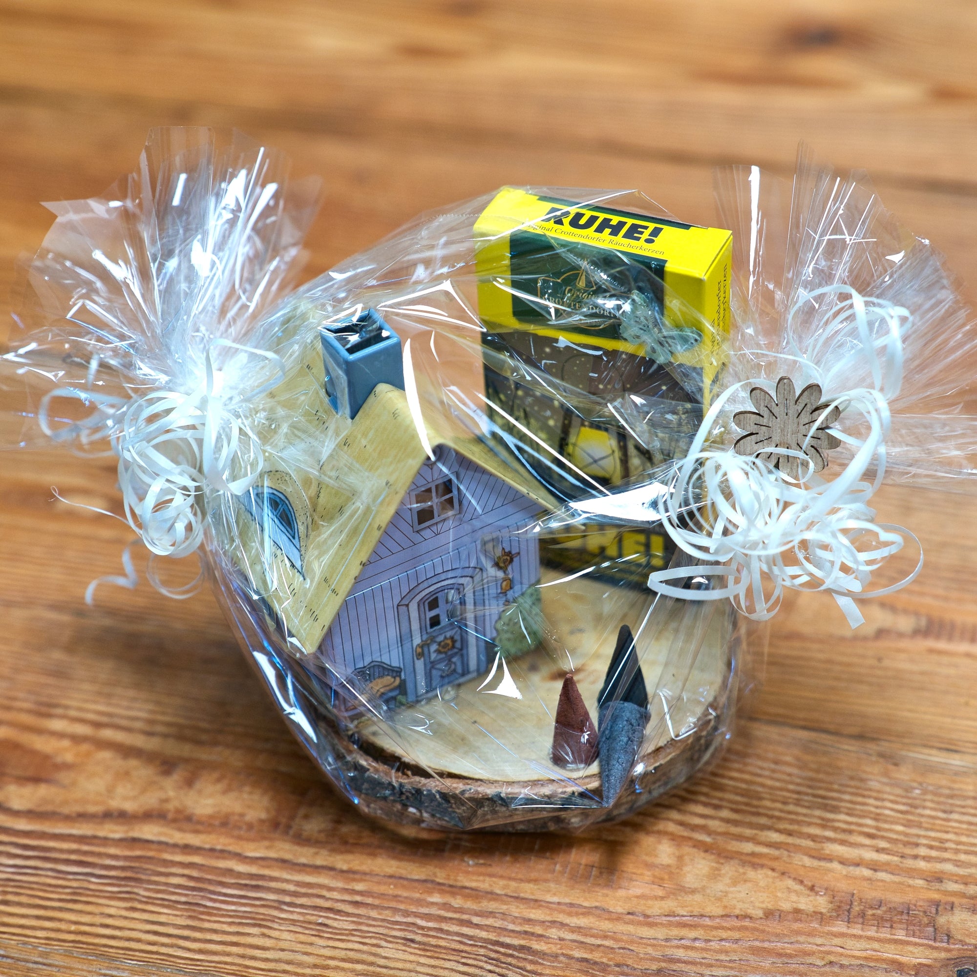 Ein in Klarsichtfolie verpackter Geschenkkorb mit weißen Bändern präsentiert das „Geschenkset Räucherhäuschen + Räucherkerzen“ von Original Crottendorfer Räucherkerzen mit Musterhaus, Gewürzflasche und Kühne-Artikel elegant arrangiert auf Holz.