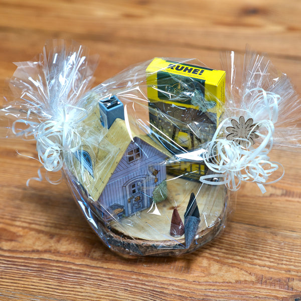 Ein in Klarsichtfolie verpackter Geschenkkorb mit weißen Bändern präsentiert das „Geschenkset Räucherhäuschen + Räucherkerzen“ von Original Crottendorfer Räucherkerzen mit Musterhaus, Gewürzflasche und Kühne-Artikel elegant arrangiert auf Holz.