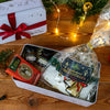 Auf einem Holztisch steht ein Original Crottendorfer Stollen Geschenkset in einer festlichen Dose, gefüllt mit Marmeladengläsern, Schokolade und Leckereien. Ein rotes Band schmückt die Dose inmitten von Lichterketten und Grünzeug und schafft so eine fröhliche Weihnachtsszene.