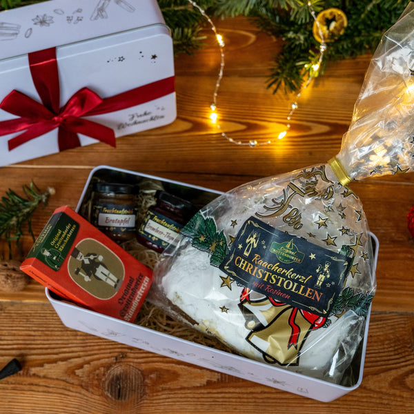 Auf einem Holztisch steht ein Original Crottendorfer Stollen Geschenkset in einer festlichen Dose, gefüllt mit Marmeladengläsern, Schokolade und Leckereien. Ein rotes Band schmückt die Dose inmitten von Lichterketten und Grünzeug und schafft so eine fröhliche Weihnachtsszene.