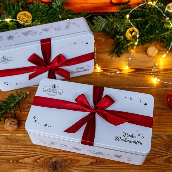 Zwei Stollen-Geschenkset-Dosen von Original Crottendorfer, verziert mit festlichen Motiven und "Frohe Weihnachten", stehen auf einer Holzfläche inmitten von Evergreens, Ornamenten, Walnüssen und Lichterketten - perfekt für Weihnachtsgeschenke.