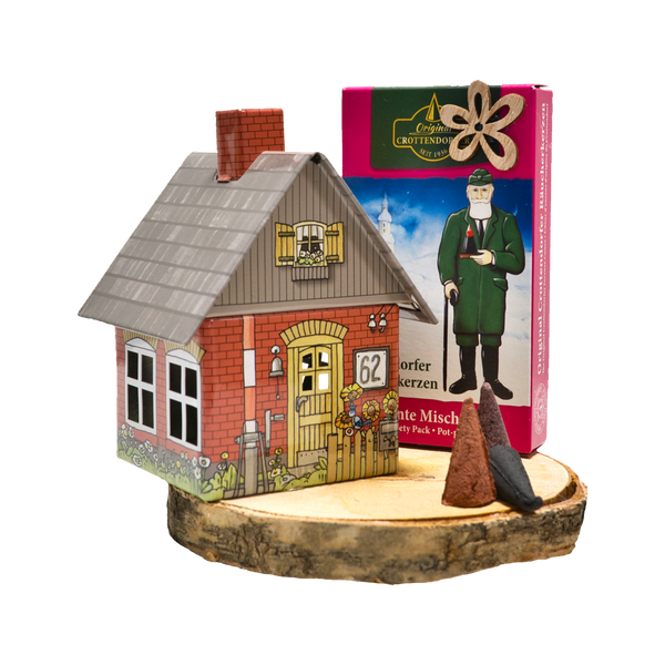 Auf einem Holzsockel mit zwei kegelförmigen Räucherkerzen sitzt ein kleines dekoratives Räucherhäuschen, das einem roten Backsteinhaus mit „62“ und einer Blumentür ähnelt. Das Set stammt aus dem Geschenkset Räucherhäuschen + Räucherkerzen von Original Crottendorfer Räucherkerzen und zeichnet sich durch seinen einzigartigen Geschenkinhalt aus.