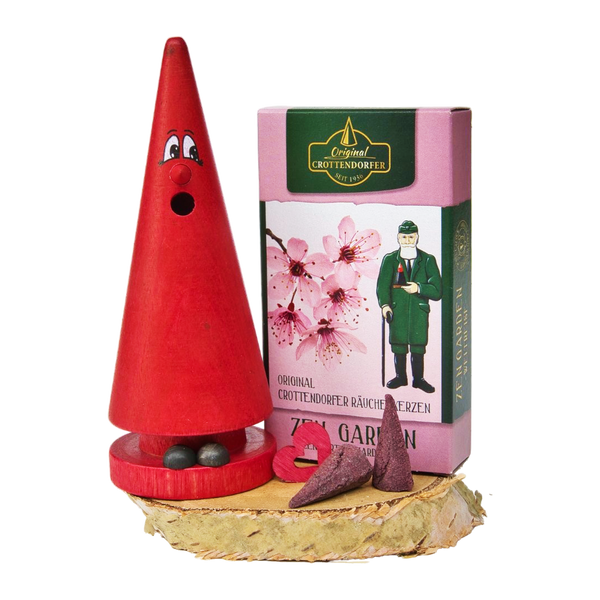 Ein roter, kegelförmiger Räucherkerzenhalter mit Augen und Mund steht auf einem Holzsockel neben Original Crottendorfer Räucherkerzen, einem herzförmigen Stück und einer rosa Blumenbox aus dem Geschenkset „Ziegenbein + Räucherkerzen“.