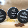 Auf einer Holzunterlage mit gelben Harzstückchen steht eine runde Dose mit der Aufschrift „Mastix in der Schraubdeckeldose“ von Original Crottendorfer Räucherkerzen, während im Hintergrund teilweise weitere Dosen mit „griechischer Räuchermischung“ zu sehen sind.