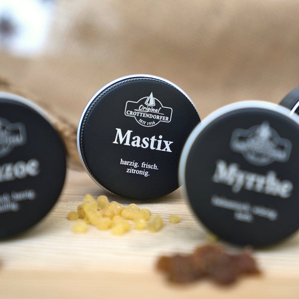 Auf einer Holzunterlage mit gelben Harzstückchen steht eine runde Dose mit der Aufschrift „Mastix in der Schraubdeckeldose“ von Original Crottendorfer Räucherkerzen, während im Hintergrund teilweise weitere Dosen mit „griechischer Räuchermischung“ zu sehen sind.