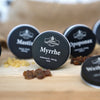 Auf einem Holzuntergrund steht eine runde schwarze Dose mit der Aufschrift „Myrrhe in der Schraubdeckeldose“ von Original Crottendorfer Räucherkerzen; im Hintergrund sind verschwommene Dosen mit Mastix und Opoponax Reinigungsweihrauch sowie Harzstücke zu erkennen.