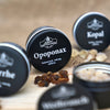 Vier schwarze Behälter mit weißen Etiketten, darunter „Oppoponax in der Schraubdeckeldose“ von Original Crottendorfer Räucherkerzen, zeigen aromatische Harzstücke auf einer Holzunterlage vor einem sanft unscharfen braunen Hintergrund.