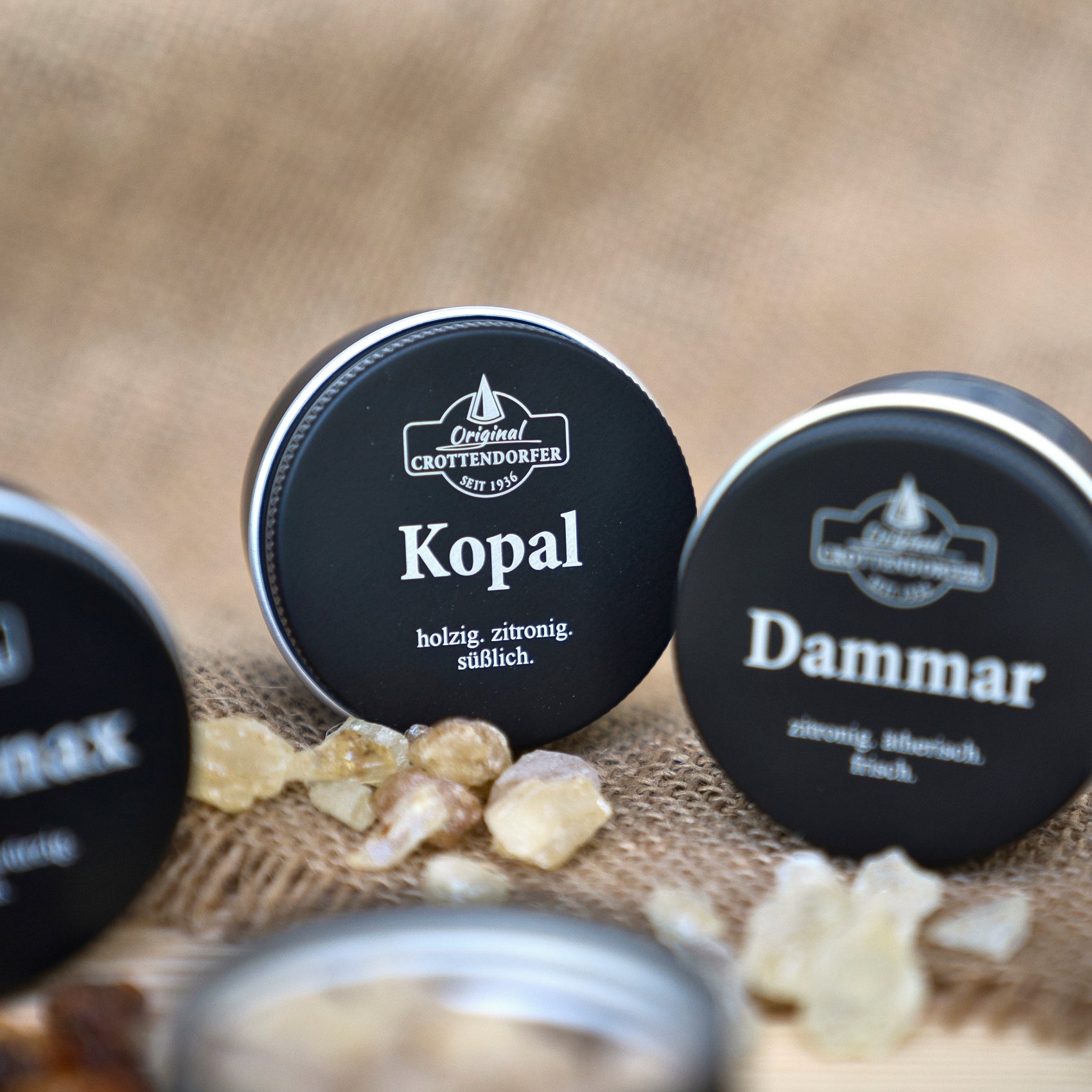 Runde schwarze Dosen mit der Aufschrift „Kopal in der Schraubdeckeldose“ von Original Crottendorfer Räucherkerzen stehen auf Jute zusammen mit kleinen Harzstückchen und verströmen holzige Zitrusdüfte sowie reinigende Eigenschaften – eine rustikale Weihrauchbrenner-Szene.