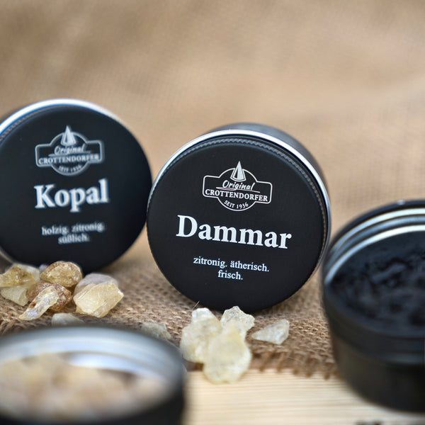 Nahaufnahme von „Dammar in der Schraubdeckeldose“ von Original Crottendorfer Räucherkerzen, umgeben von gelblichen Harzstückchen und einer Blechdose mit dunklem Inhalt auf einer natürlich strukturierten Oberfläche, die einen ätherischen Duft verströmt.