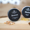 Auf einem Holzstück steht eine runde schwarze Dose mit der Aufschrift „Benzoe in der Schraubdeckeldose“ von Original Crottendorfer Räucherkerzen, umgeben von Harzstücken. Eine weitere ähnliche Dose und Harz sind im Hintergrund verschwommen zu sehen und lassen einen dezenten Vanilleduft erahnen.