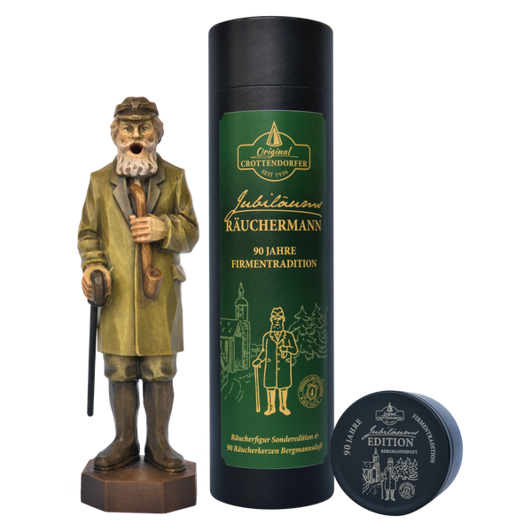 Ein hölzerner Jubiläums Räuchermann von Original Crottendorfer Räucherkerzen steht neben seiner großen dunkelgrünen Geschenkdose und einer kleinen runden Dose, die alle mit klassischen gold-weißen Motiven und Illustrationen versehen sind.