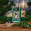 Eine „Original Crottendorfer Räucherkerzen“ Räucherkerzen Jubiläums Edition Tannenduft-Box steht auf einer Holzoberfläche mit immergrünen Zweigen, begleitet von zwei dunkelgrünen Räucherkegeln aus ätherischen Tannenharzen, die den Waldduft vor einem verschwommenen Hintergrund verstärken.