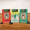 Drei Schachteln Räucherkerzen Jubiläumsedition Tannenduft von Original Crottendorfer Räucherkerzen stehen auf einer Holzoberfläche. Die Verpackung ist in Rot, Grün und Blaugrün gehalten und mit traditionellen deutschen Weihnachtsillustrationen versehen. Um sie herum sind kleine schwarze Räucherkegel mit ätherischem Tannenharz verstreut.