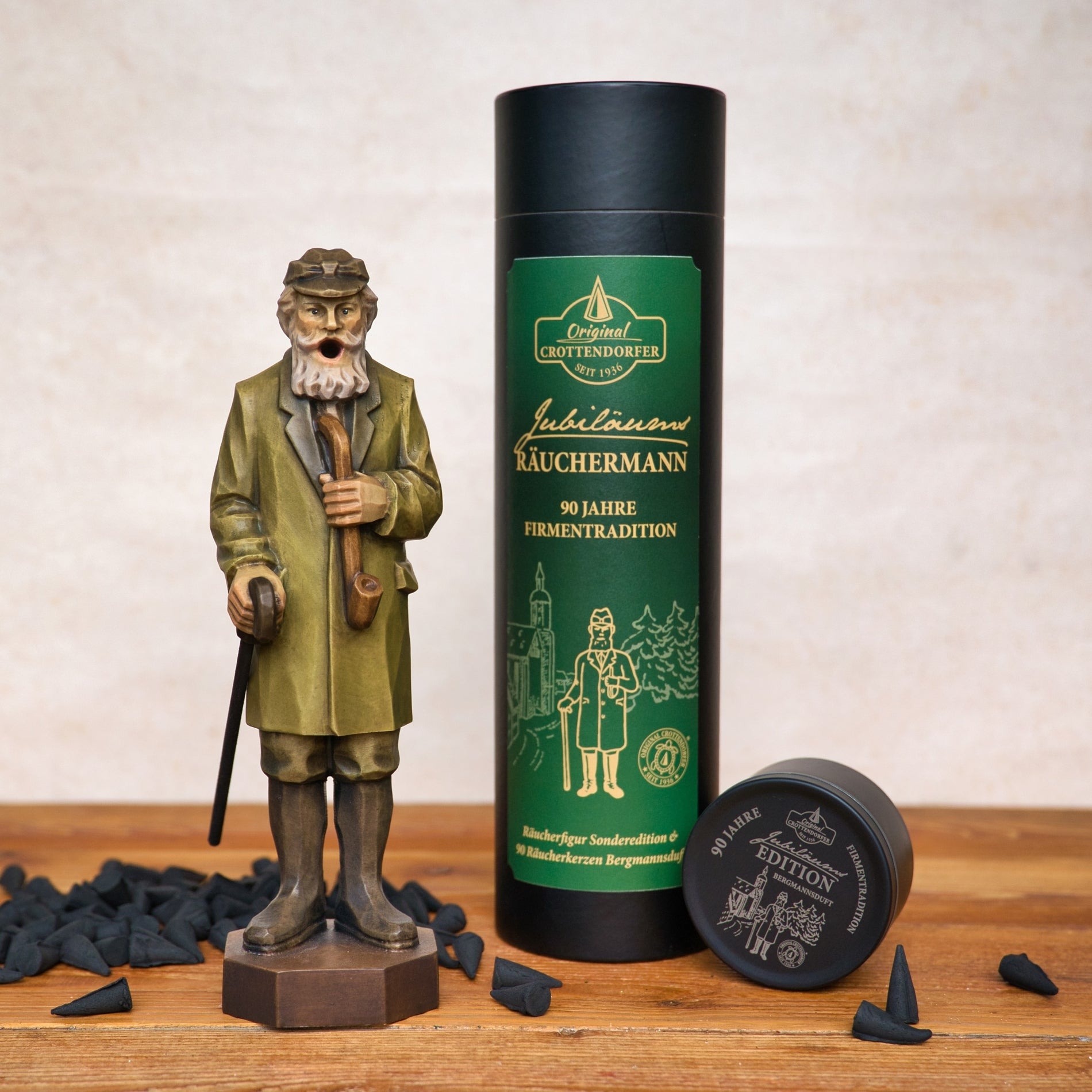 Der Jubiläums Räuchermann in der schwarzen Geschenkdose von Original Crottendorfer Räucherkerzen steht neben einer grün-schwarzen Räucherpackung und einem kleinen Behälter, mit auf der Holzoberfläche verstreuten Räucherkegeln.