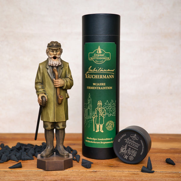 Der Jubiläums Räuchermann in der schwarzen Geschenkdose von Original Crottendorfer Räucherkerzen steht neben einer grün-schwarzen Räucherpackung und einem kleinen Behälter, mit auf der Holzoberfläche verstreuten Räucherkegeln.