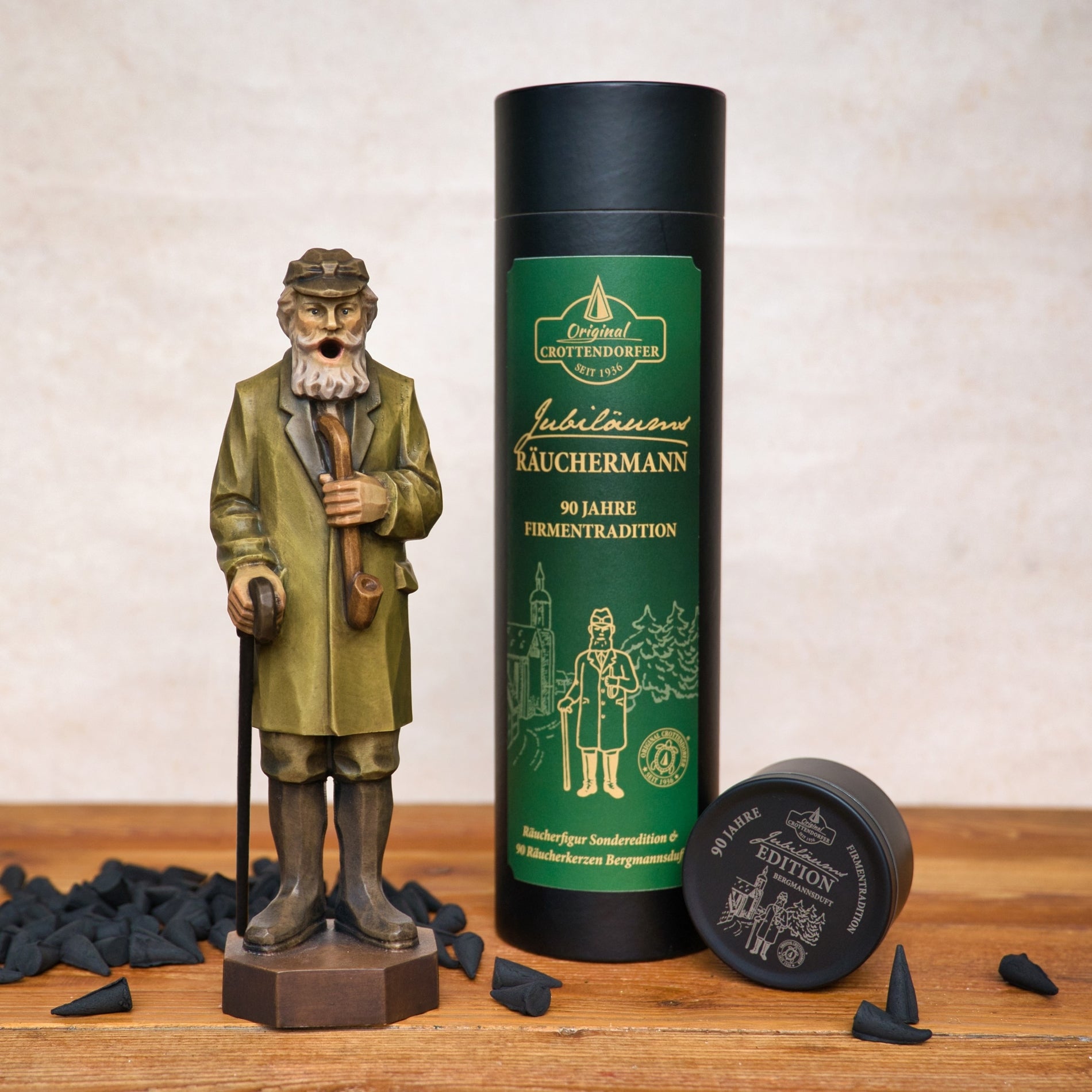 Der Jubiläums Räuchermann in der schwarzen Geschenkdose von Original Crottendorfer Räucherkerzen steht neben einer grün-schwarzen Räucherpackung und einem kleinen Behälter, mit auf der Holzoberfläche verstreuten Räucherkegeln.