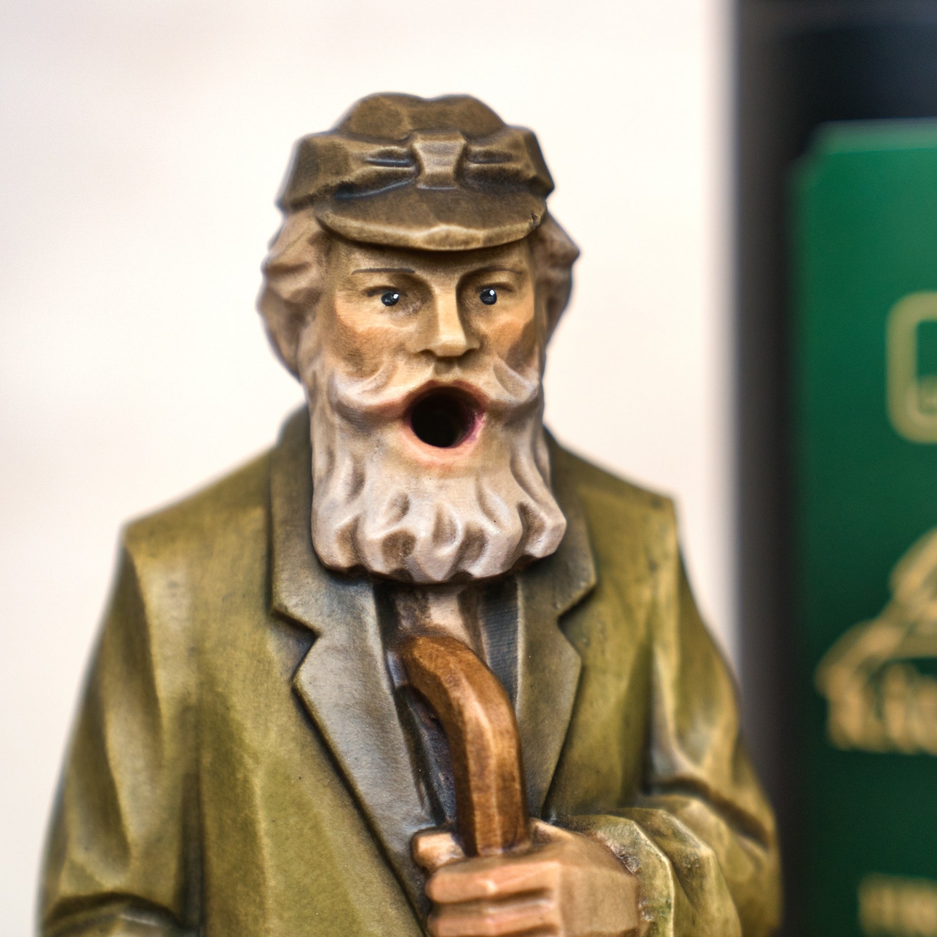 Eine Nahaufnahme des Jubiläums Räuchermann in der schwarzen Geschenkdose von Original Crottendorfer Räucherkerzen zeigt einen älteren Mann mit grauem Bart, Mütze und Gehstock. Im Hintergrund ist teilweise ein grün-schwarzer Gegenstand zu sehen.