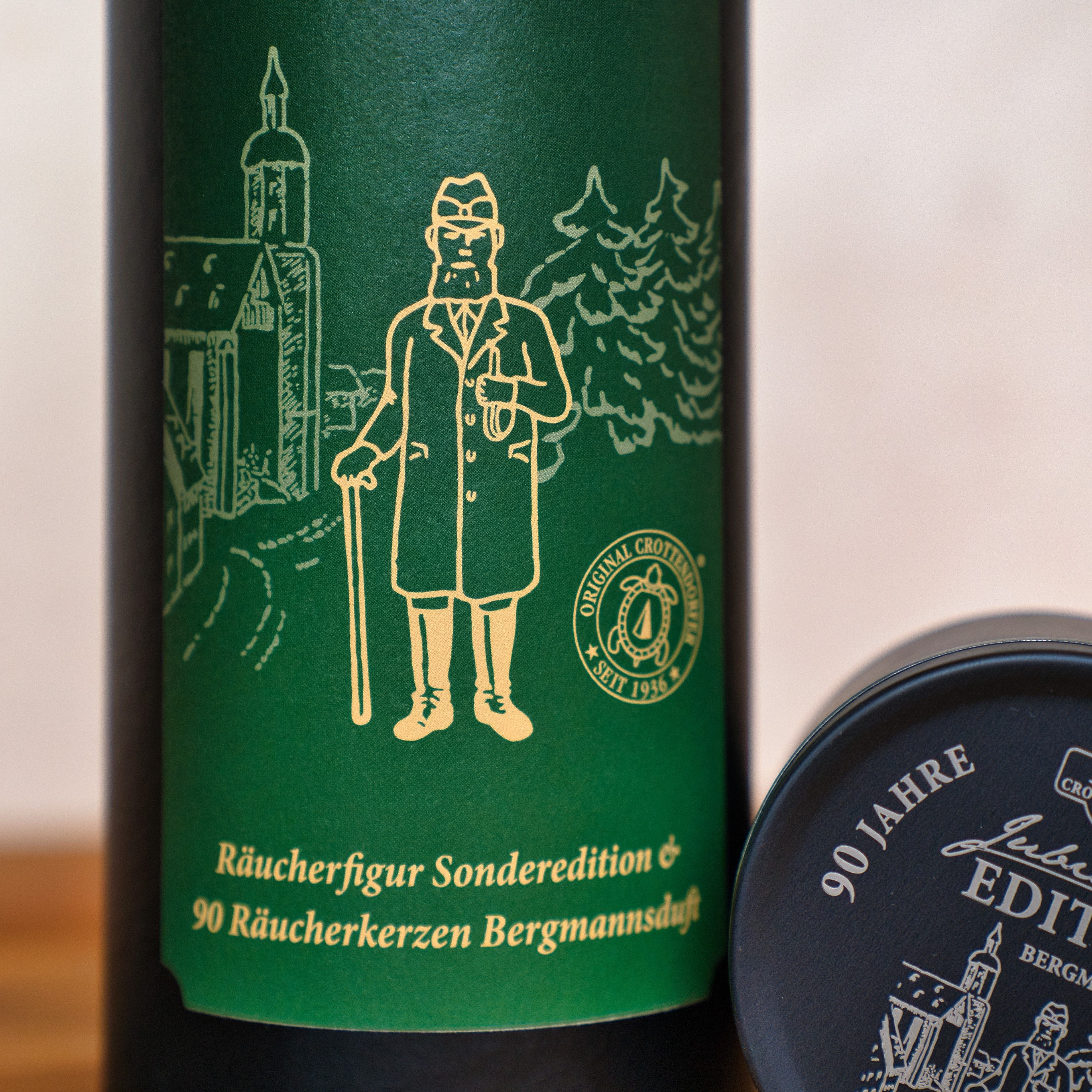 Eine Nahaufnahme des Etiketts Jubiläums Räuchermann von Original Crottendorfer Räucherkerzen zeigt einen illustrierten Mann mit Stock und Pfeife im Stil der erzgebirgischen Handwerkskunst vor Gebäuden mit deutschem Text und Wappen. Ein runder schwarzer Geschenkdosendeckel ist sichtbar.