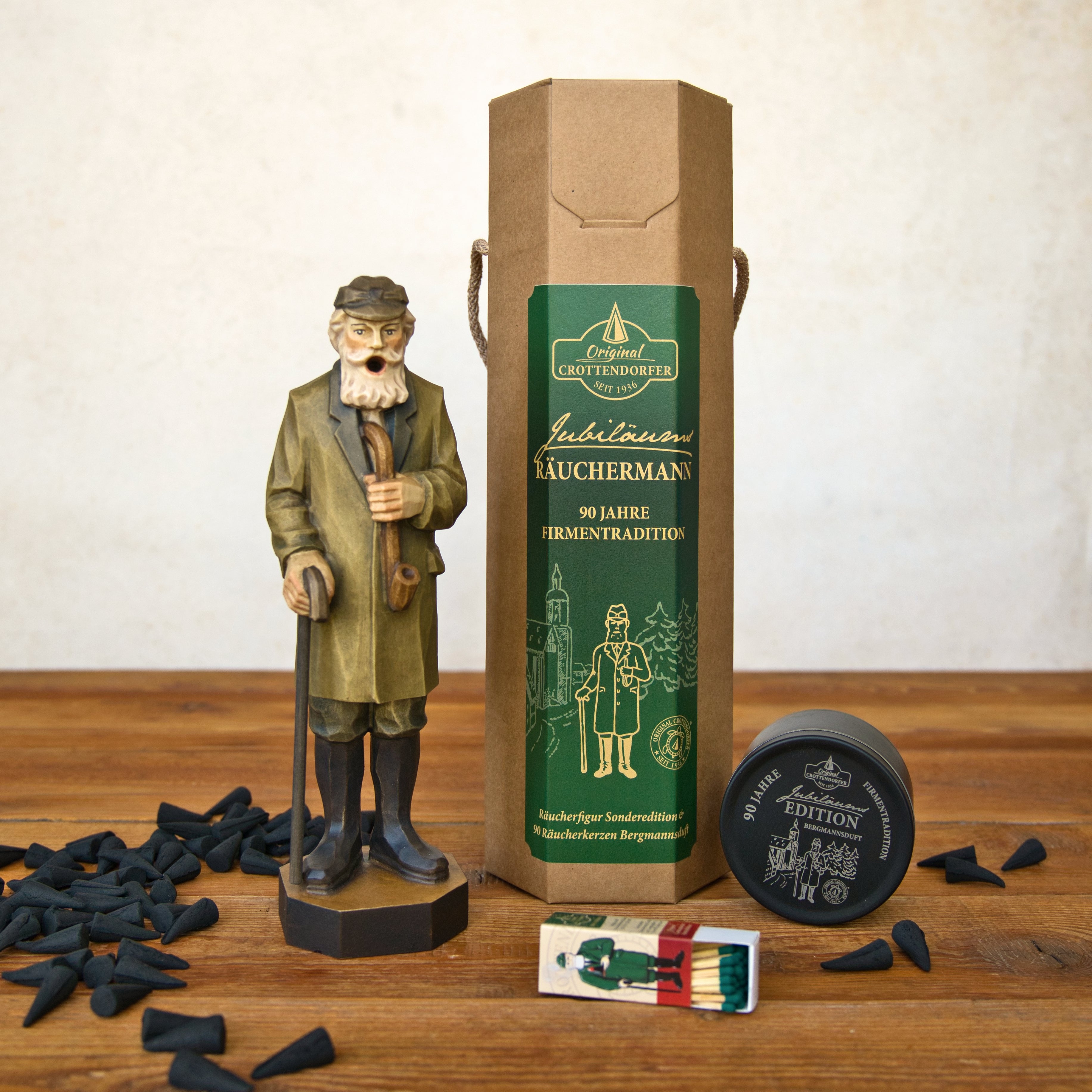 Eine Original-Crottendorfer Räucherkerze „Jubiläums-Räuchermann“ in einer Geschenkverpackung, zusammen mit einer braunen Pappschachtel, schwarzen „Bergmannsduft“-Räucherkerzenkegeln, einer kleinen runden Blechdose und einer Streichholzschachtel, ist auf einer Holzunterlage angeordnet. Die Verpackung ist mit einem grünen Etikett mit gold-weißer Beschriftung versehen, das die erzgebirgische Tradition widerspiegelt.