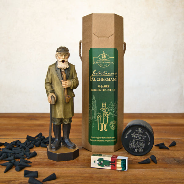 Eine Original-Crottendorfer Räucherkerze „Jubiläums-Räuchermann“ in einer Geschenkverpackung, zusammen mit einer braunen Pappschachtel, schwarzen „Bergmannsduft“-Räucherkerzenkegeln, einer kleinen runden Blechdose und einer Streichholzschachtel, ist auf einer Holzunterlage angeordnet. Die Verpackung ist mit einem grünen Etikett mit gold-weißer Beschriftung versehen, das die erzgebirgische Tradition widerspiegelt.