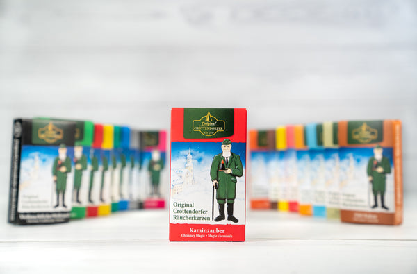 Eine Schachtel Räucherkerzen Kaminzauber von Original Crottendorfer Räucherkerzen ist mit einer Trachtenabbildung auf der Verpackung versehen. Der Zimtduft weckt Nostalgie, während ähnliche Schachteln im Hintergrund unscharf sind.