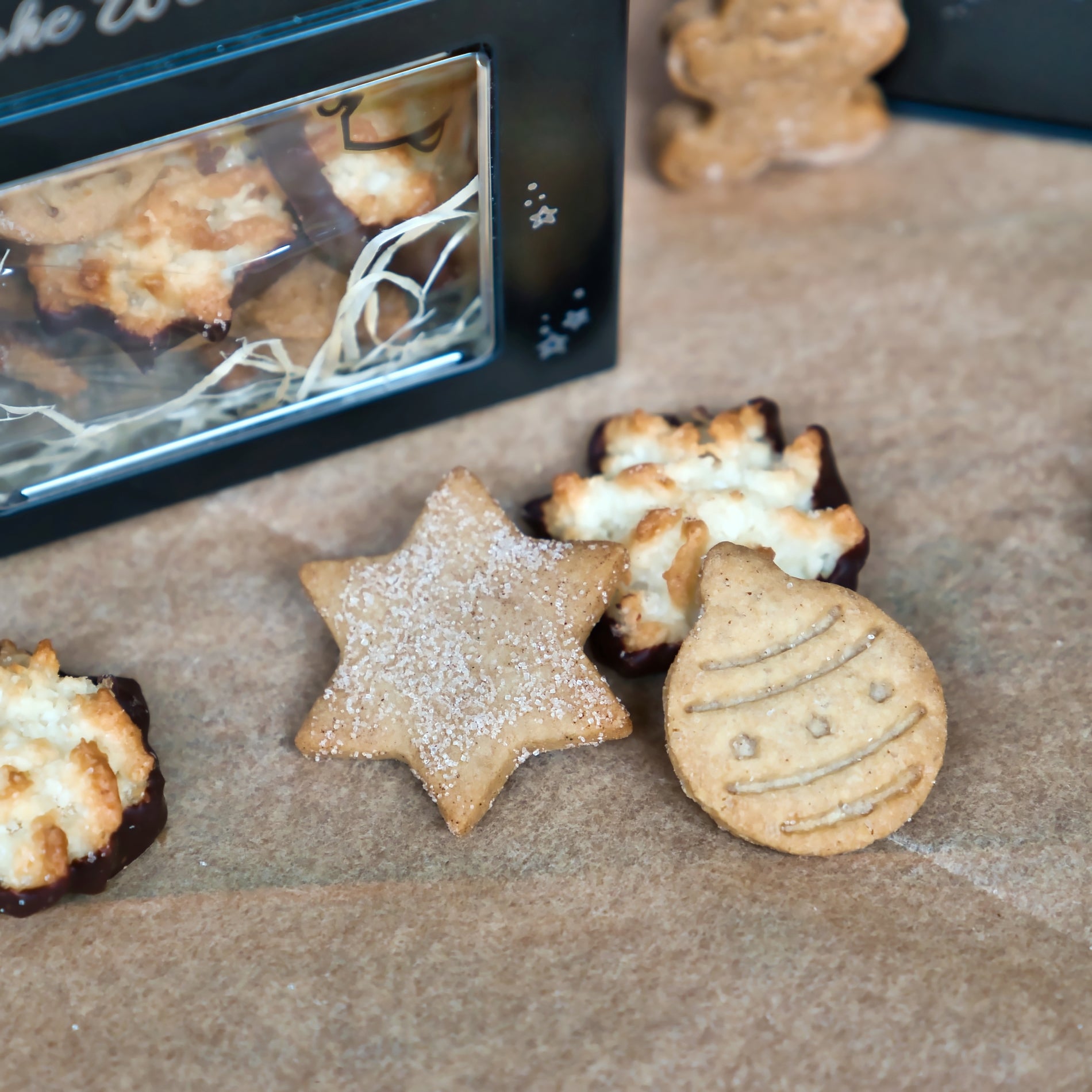 Verschiedene Weihnachtskekse in Form von Sternen und Ornamenten sowie schokoladige Leckereien werden mit einer geschenkfertigen Original Crottendorfer Gebäckmischung in der festlichen schwarzen Keksdose im Hintergrund präsentiert.