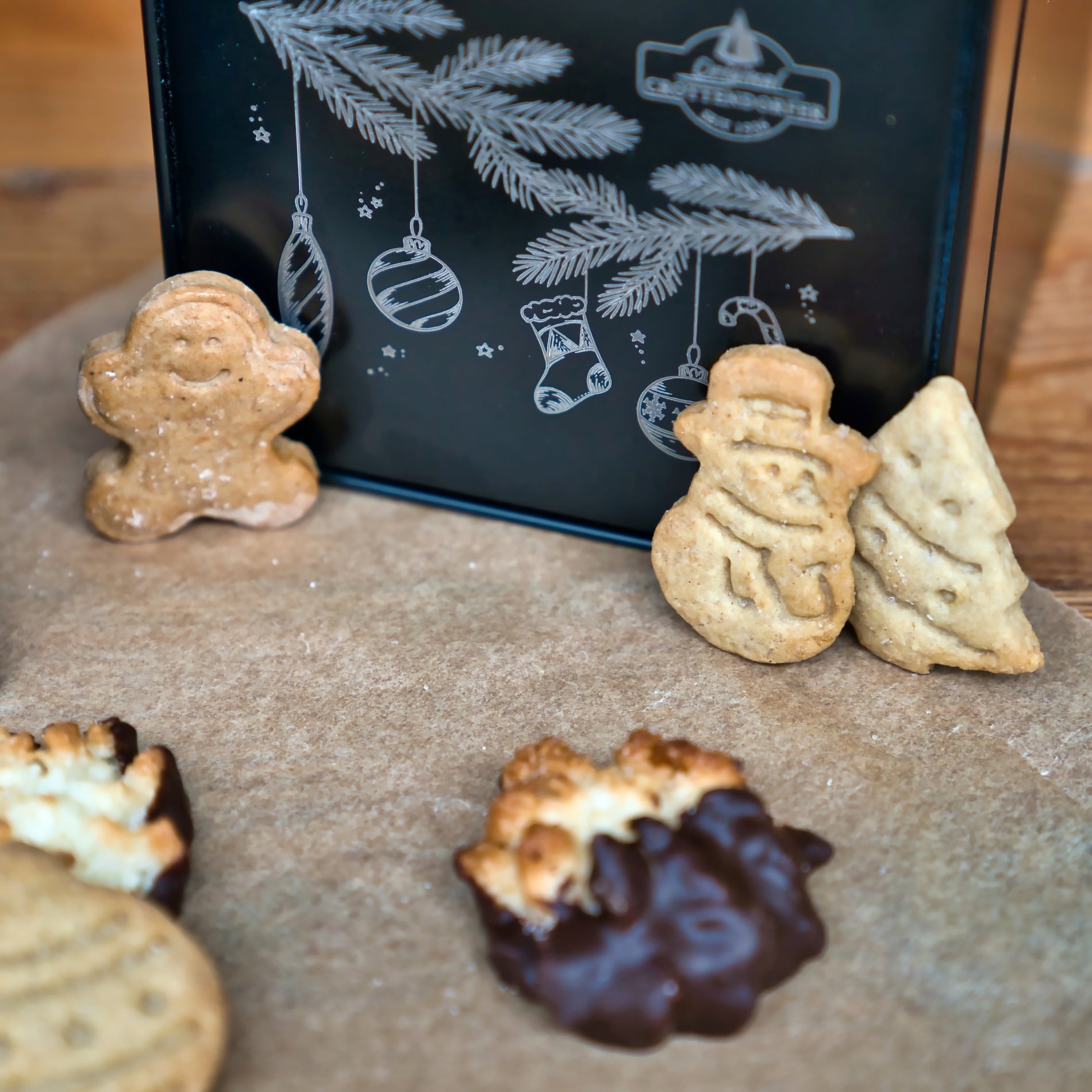 Verschiedene Weihnachtsplätzchen - Lebkuchenmann, Schneemann und Baum - sind auf Pergament vor einer Original Crottendorfer Keksdose, schwarz, arrangiert und mit festlichem weißen Weihnachtsschmuck verziert.