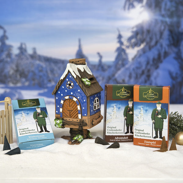 Das Original Crottendorfer Räucherhaus aus Keramik, inklusive 3 Packungen Räucherkerzen – Winter, Advent, Zimt – steht auf Kunstschnee mit Tannen und verschneitem Hintergrund und sorgt so für eine stimmungsvolle weihnachtliche Dekoration.