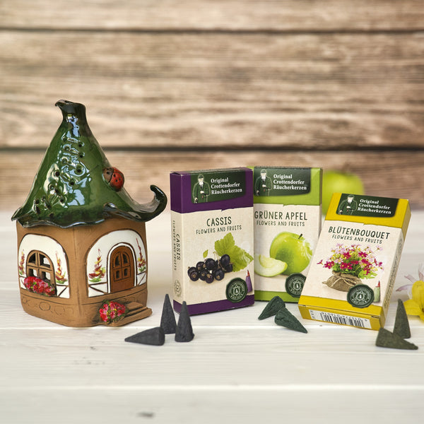 Ein originales Crottendorfer Räucherhaus aus Keramik, inklusive 3 Packungen Räucherkerzen (schwarze Johannisbeere, grüner Apfel, Blumenbouquet), steht auf einer Holzunterlage vor einem unscharfen Holzhintergrund.