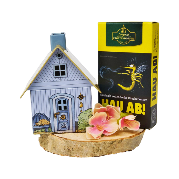 Das Geschenkset „Räucherhäuschen + Räucherkerzen“ von Original Crottendorfer Räucherkerzen besteht aus einem dekorativen Räucherhalter auf einem Holzsockel, rosa Blumen und einer gelben „Hau Ab!“-Schachtel mit Mückenmotiv – ideal als Geschenk.