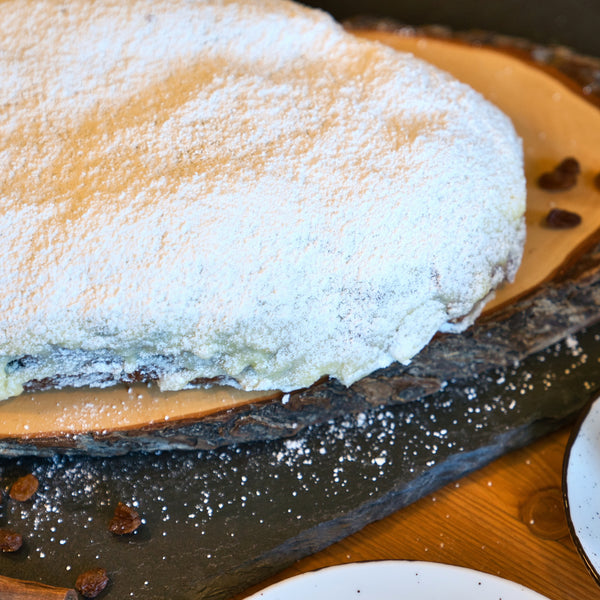 Ein Stück Original Crottendorfer Raacherkerzl-Christstollen mit Rosinen, großzügig mit Puderzucker bestäubt, ruht auf einer rustikalen Holzplatte. Auf dem dunklen Schiefer daneben sind Rosinen und festliche Zutaten verstreut.