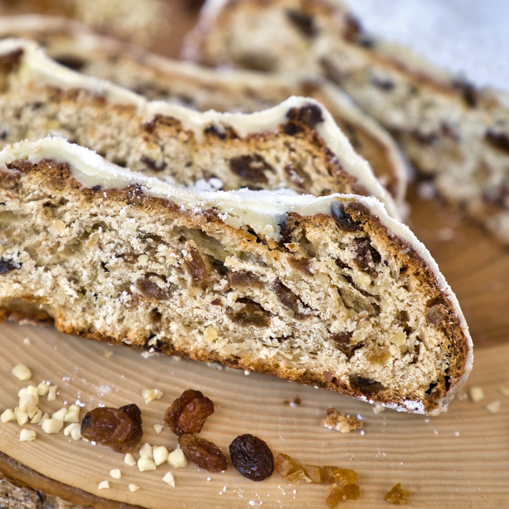Drei Scheiben Original Crottendorfer Raacherkerzl-Christstollen mit Rosinen, dicht mit sichtbaren Trockenfrüchten und Nüssen bestückt, liegen auf einer Holzunterlage mit gestreuten Rosinen und gehackten Nüssen und erinnern an klassisches Stollen-Weihnachtsgebäck.