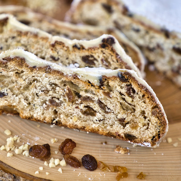 Drei Scheiben Original Crottendorfer Raacherkerzl-Christstollen mit Rosinen, dicht mit sichtbaren Trockenfrüchten und Nüssen bestückt, liegen auf einer Holzunterlage mit gestreuten Rosinen und gehackten Nüssen und erinnern an klassisches Stollen-Weihnachtsgebäck.