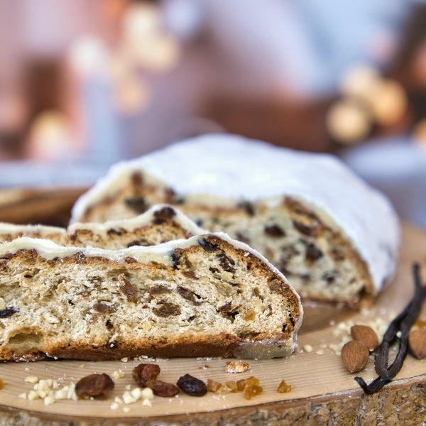 Eine Nahaufnahme von Original Crottendorfer Raacherkerzl-Christstollen mit Rosinen, bestäubt mit Puderzucker und mit Rosinen im Inneren, auf einem Holzbrett ruhend, mit verschwommenen festlichen Lichtern im Hintergrund.