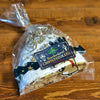Ein Raacherkerzl-Christstollen mit Rosinen von Original Crottendorfer, bestäubt mit Puderzucker, liegt auf einer Holzplatte. Die durchsichtige Verpackung ist mit festlichen Gold- und Marinemotiven verziert und weist auf die lange Haltbarkeit hin.