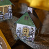 Ein kleines Räucherhäuschen „Huthaus“ von Original Crottendorfer Räucherkerzen, dessen Papierwände mit illustrierten Figuren verziert sind, die neben der Tür sitzen und stehen, thront auf Felsen. Sein grünes Dach und die Steinakzente unterstreichen seinen urigen Charme, während dezente metallische Akzente an den Charme eines traditionellen Räucherkerzenhalters erinnern.