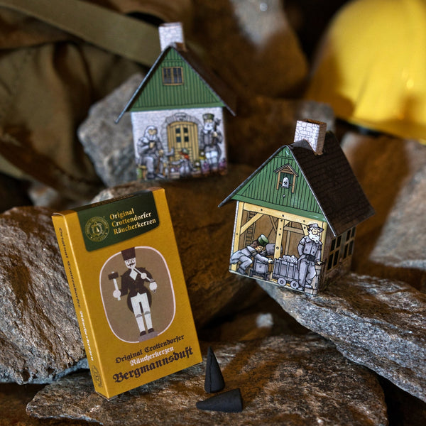 Zwei Modellhäuschen des Modells „Räucherhäuschen Huthaus“ von Original Crottendorfer Räucherkerzen, jeweils mit abgebildeten Bergleuten, sind auf grauen Felsen neben einer gelben „Bergmannsduft“-Räucherkegel-Schachtel und zwei schwarzen Räucherkegelhaltern zu sehen. Im Hintergrund ist ein gelbes Objekt verschwommen zu erkennen.