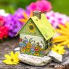 Das Räucherhäuschen Blumenmeer von Original Crottendorfer Räucherkerzen ist ein kleines dekoratives Metallhaus mit grünem Dach und aufgemalten Blumen, perfekt als skurriler Garten-Räucherkerzenhalter inmitten lebendiger Blüten.