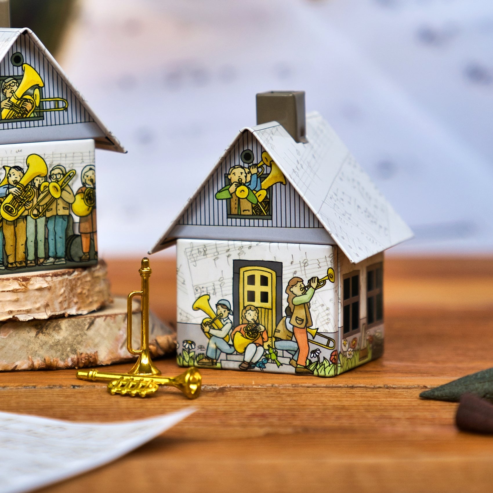 Das Räucherhäuschen Posaunenchor von Original Crottendorfer Räucherkerzen ist ein kleines dekoratives Häuschen mit Messingmusikanten, einer kleinen goldenen Trompete davor und einem weiteren ähnlichen Häuschen, das teilweise auf der linken Seite zu sehen ist - alles auf einer Holzfläche.