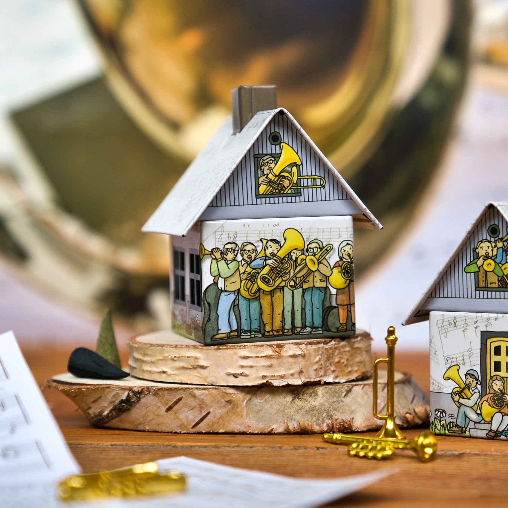 Das Räucherhäuschen Posaunenchor von Original Crottendorfer Räucherkerzen ist ein kleines Modellhaus mit Blaskapellen-Illustrationen, auf Holz gesetzt, mit Miniatur-Metalltrompete und -horn daneben und verschwommenen Blechblasinstrumenten im Hintergrund.