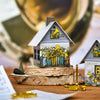 Das Räucherhäuschen Posaunenchor von Original Crottendorfer Räucherkerzen ist ein kleines Modellhaus mit Blaskapellen-Illustrationen, auf Holz gesetzt, mit Miniatur-Metalltrompete und -horn daneben und verschwommenen Blechblasinstrumenten im Hintergrund.