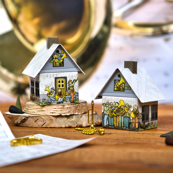 Das Räucherhäuschen Posaunenchor von Original Crottendorfer Räucherkerzen besteht aus zwei kleinen Papierhäuschen, die mit Musikern aus Messing geschmückt sind. Sie stehen auf Birkenrindenholz und sind mit Metalltrompeten und musikalischen Accessoires bestückt.