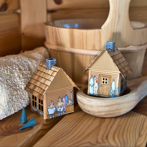 Zwei kleine Räucherhäuschen-Sauna-Modelle aus Holz von Original Crottendorfer Räucherkerzen, die jeweils mit einer illustrierten Saunaszene verziert sind, stehen auf einer Holzunterlage neben einem beigen Handtuch, einem Holzeimer, einer Saunakelle und zwei kegelförmigen blauen Räucherkerzenhaltern.