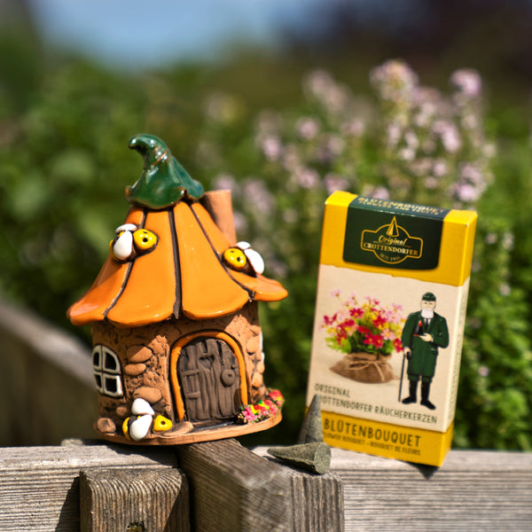 Ein kleines, verspieltes „Original Crottendorfer Räucherhaus Keramik Bienenhaus“ mit grünem Dach steht liebevoll auf einer Holzleiste neben einer gelben Schachtel mit Räucherkegeln und einem zierlichen Messingteller, im Hintergrund ist verschwommenes Grün zu sehen.