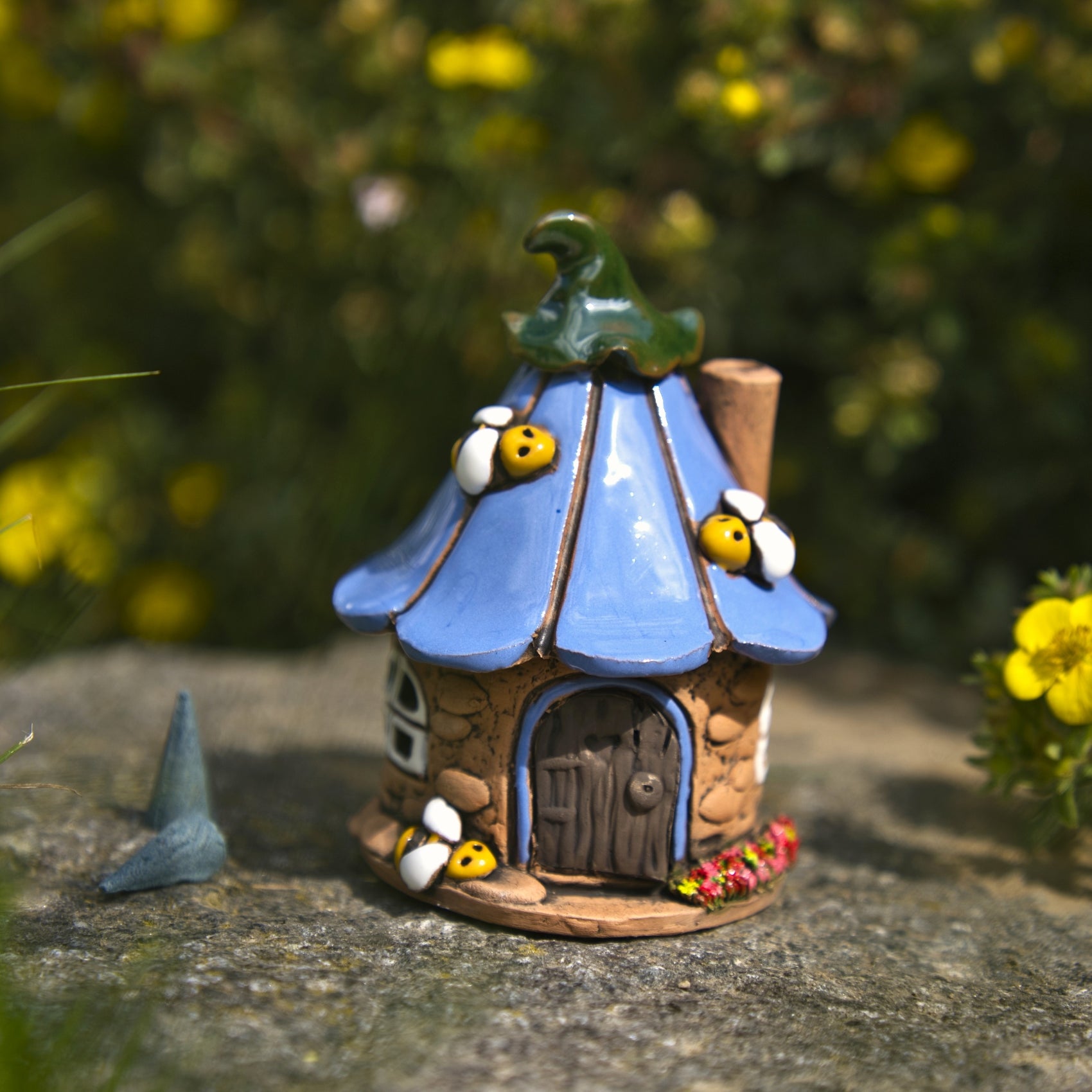 Ein bezauberndes „Räucherhaus“-Keramik-Bienenhaus von Original Crottendorfer mit blauem Dach, grünem Schornstein und entzückenden Bienenverzierungen steht liebevoll hergestellt auf einer Steinunterlage, umgeben von Grünpflanzen und gelben Blumen.