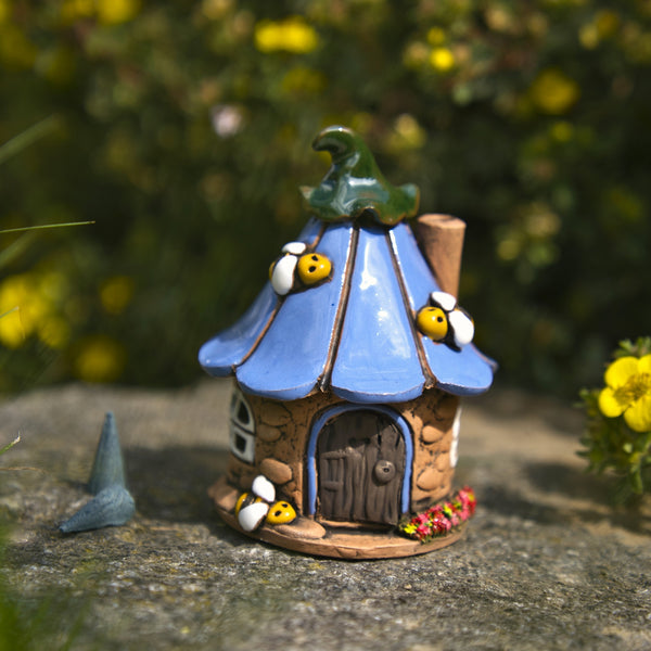 Ein bezauberndes „Räucherhaus“-Keramik-Bienenhaus von Original Crottendorfer mit blauem Dach, grünem Schornstein und entzückenden Bienenverzierungen steht liebevoll hergestellt auf einer Steinunterlage, umgeben von Grünpflanzen und gelben Blumen.